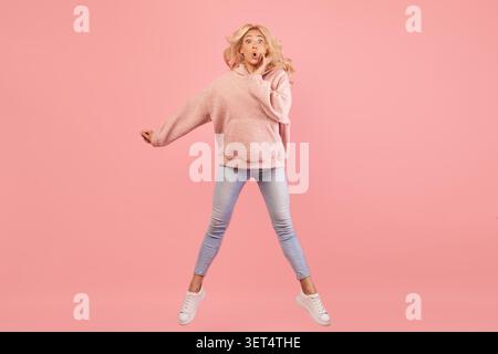Sorpresa giovane donna che salta in Joy con le mani sul viso, sfondo rosa, concetto Happy Emotion Foto Stock