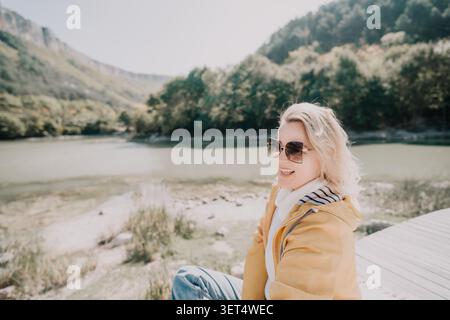 Donna sorridente in occhiali da sole seduta accanto a un lago Foto Stock