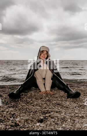 Donna spiaggia invernale giovane donna che indossa un cappello ushanka e una giacca in pelle si siede su una spiaggia di ciottoli sotto un cielo nuvoloso Foto Stock