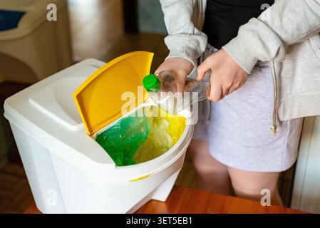 Primo piano di una donna che mette una bottiglia di plastica vuota in un bidone giallo a casa Foto Stock