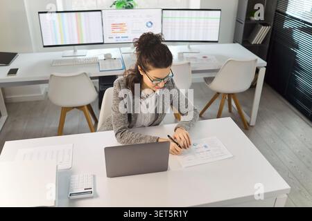 Analista aziendale che lavora su notebook con grafici, documenti cartacei e dati finanziari in ufficio Foto Stock