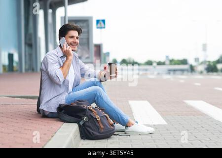 Uomo che aspetta fuori dall'aeroporto, bevendo caffè e parlando al cellulare Foto Stock