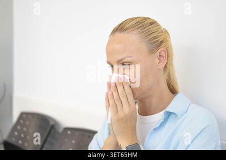 Donna seduta in una sala d'attesa, soffiando il naso in un fazzoletto di carta, indicando che si sente male a causa di un raffreddore, influenza, o allergie stagionali, exper Foto Stock