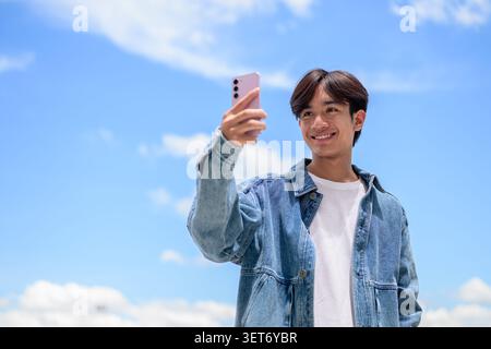 Fotografia orizzontale di uno stile di vita dall'angolazione bassa di un bel giovane tailandese del sud-est asiatico Isan non-binario che sorride felicemente mentre tiene sollevato il suo smartphone Foto Stock
