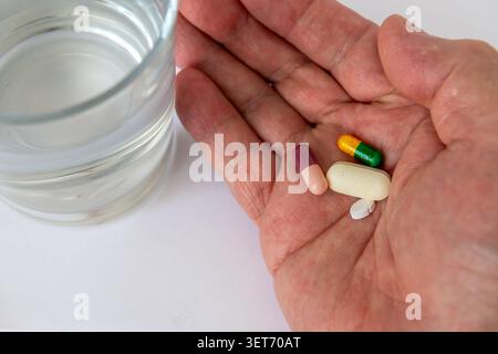 Compresse e capsule di medicina nel palmo di una mano con un bicchiere d'acqua al fianco, isolato su bianco. Foto Stock