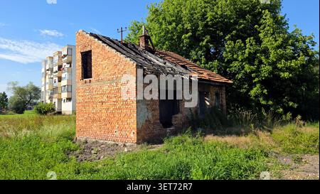 Un'ampia immagine di un cottage in mattoni a un piano in rovina con un tetto parzialmente distrutto in piastrelle di argilla seduto in un campo erboso con un contemporaneo multipiano Foto Stock