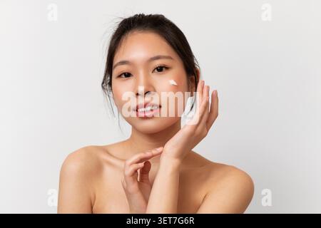 Giovane donna asiatica che applica la crema per la pelle sul viso a casa, concetto per la cura della pelle Foto Stock