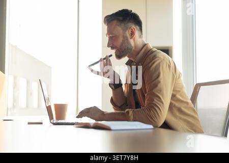 Uomo caucasico che lavora da casa, utilizzando laptop e registratore vocale in impostazione ufficio Foto Stock