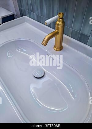 Un lavandino nuovo si trova su un ripiano del bagno con un rubinetto dorato. Il rubinetto ha un manico bianco che offre un look moderno. È pronto per l'uso quotidiano Foto Stock