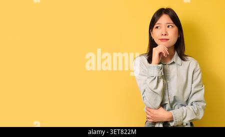 Donna asiatica premurosa che guarda in alto con la mano sul mento, sfondo giallo, concetto di perdita di peso Foto Stock