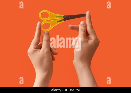 Mani per bambini che tengono in mano le forbici di sicurezza gialle con contrassegni righello su uno sfondo arancione brillante, lo sfondo arancione brillante fornisce un vivace Co Foto Stock