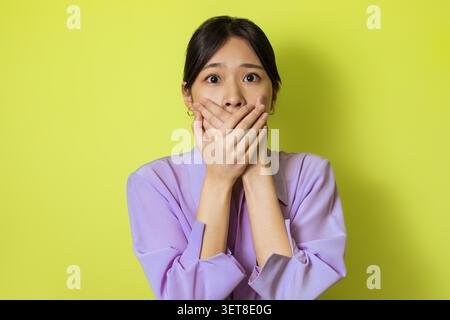 Sorpresa giovane donna asiatica che copre bocca con le mani, sfondo verde Foto Stock