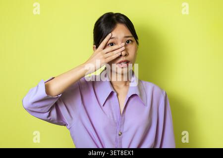 Preoccupata giovane donna asiatica che copre un occhio con la mano, sfondo verde, concetto di benessere mentale Foto Stock