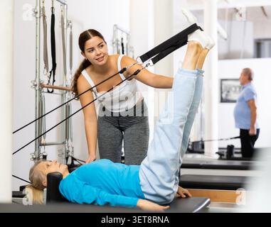 Cura allenatore donna che controlla e aiuta donna anziana che si esercita su Pilates reformer macchina in centro di salute Foto Stock