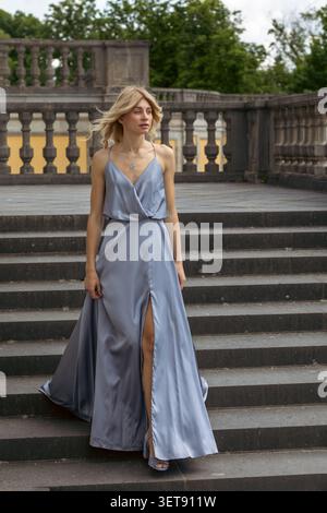 Una modella cammina lungo gradini di pietra indossando un abito blu. Ha i capelli lunghi e ci sono alberi sullo sfondo. L'impostazione è una posizione all'aperto duraturo Foto Stock