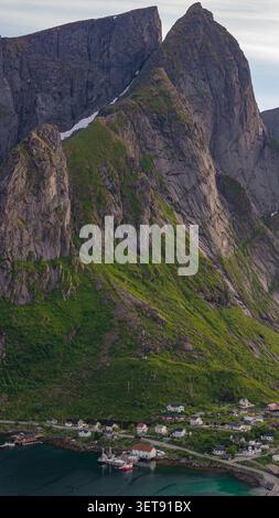 Le ripide cime rocciose sovrastano un pittoresco villaggio costiero norvegese annidato dalle acque turchesi di Lofoten. Scogliere coperte di muschio e. Foto Stock