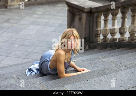 Una donna con lunghi capelli biondi siede sulle scale di un edificio storico. Tiene un bicchiere mentre guarda la telecamera. L'ambiente è luminoso e luminoso Foto Stock