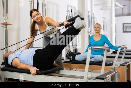 Giovane istruttrice positiva che controlla i movimenti dell'uomo anziano che si allunga sulle attrezzature del riformatore Pilates in studio Foto Stock