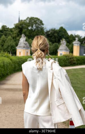 Una donna cammina su un sentiero in un giardino. Ha un blazer sulle spalle. Sullo sfondo ci sono alberi ed edifici storici. Il cielo è C. Foto Stock