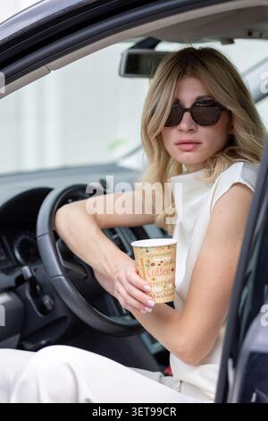 Una donna è seduta sul sedile di guida di un'auto, tenendo in mano una tazza di caffè. Sta indossando gli occhiali da sole e guardando fuori. Lo sfondo viene visualizzato Foto Stock