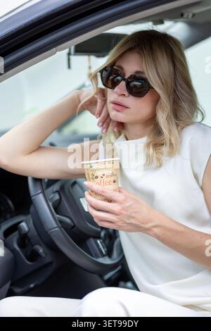 Una donna siede dentro una macchina con la porta aperta. Tiene una tazza di caffè in una mano e sembra che stia pensando. Il sole splende e sembra rilassata Foto Stock