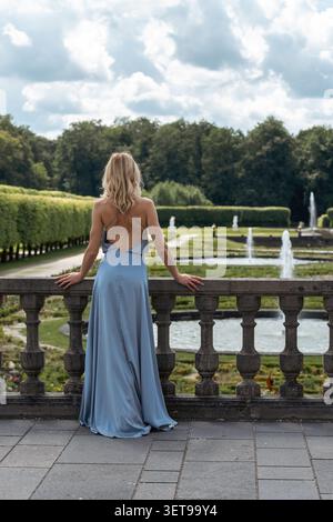 Una donna con un vestito leggero si appoggia contro una ringhiera di pietra. Si affaccia su un grande giardino con fontane, alberi e un cielo ampio. E' estate e l'ar Foto Stock
