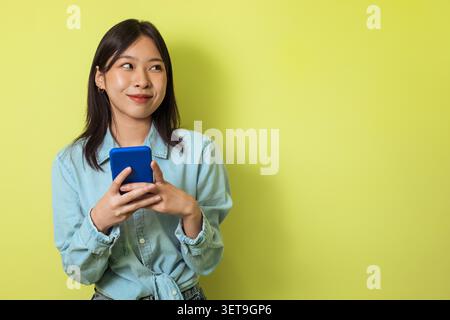 Felice donna asiatica che tiene in mano lo smartphone premuroso, sfondo giallo, concetto tecnologico Foto Stock