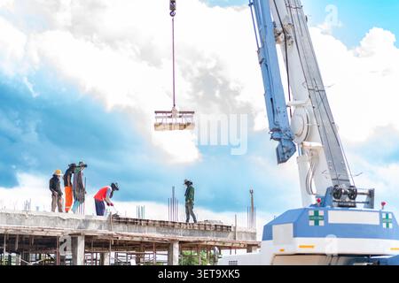 Team di costruzione che gestisce un carico pesante sospeso da un cavo di gru su una struttura dell'edificio sotto un cielo nuvoloso e luminoso. Foto Stock