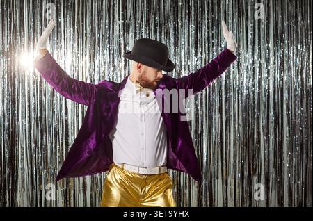 Uomo simpatico e simpatico che indossa giacca viola, pantaloni dorati, cappello, guanti che ballano energicamente in performance Foto Stock
