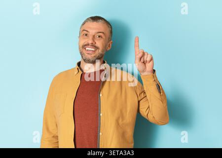 Sorridente uomo caucasico di mezza età che punta verso l'alto su spazio copia, sfondo azzurro Foto Stock