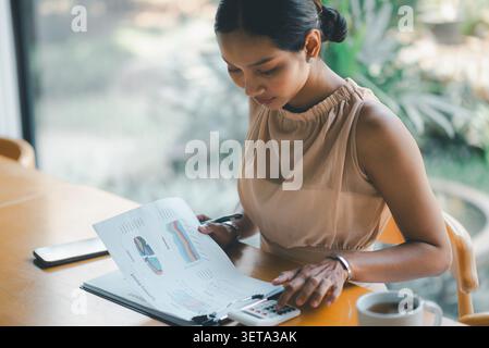 Donna d'affari asiatica che esamina i report finanziari con calcolatrice, grafici e caffè in un moderno spazio di lavoro, focalizzata sulla contabilità, la pianificazione del budget, i Foto Stock