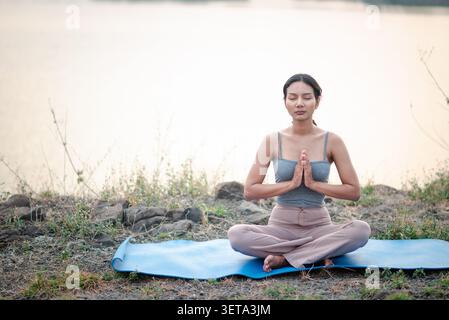 Giovane donna asiatica che medita a gambe incrociate su un tappetino blu da yoga accanto a un lago tranquillo, mani in preghiera, mostrando consapevolezza, respirazione, benessere all'aperto Foto Stock