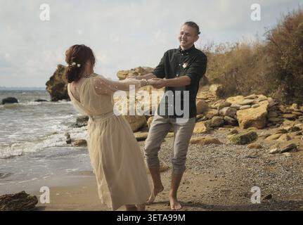 Coppia romantica e sorridente che danzano in riva al mare. Stanno girando a piedi nudi sulla riva del mare. La coppia si sta tenendo per mano. Stanno sorridendo e lau Foto Stock