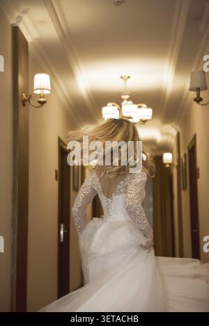 Splendida sposa bionda in abito lungo bianco che corre nel corridoio dell'hotel, abito alla moda con vista posteriore con corsetto e spalle aperte. Capelli lisci e. Foto Stock