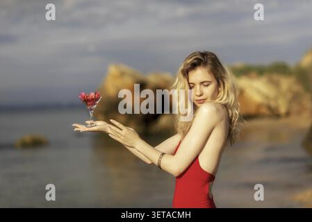 Snella bionda in un costume da bagno rosso si erge su una spiaggia di sabbia, tenendo un bicchiere di fiori, si gode il sole in una splendida giornata in spiaggia. Giovane donna p Foto Stock