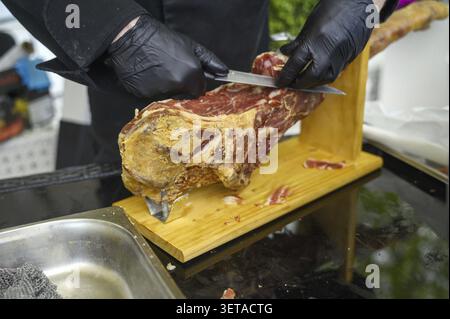 Primo piano di un venditore che affetta a mano pezzi freschi di prosciutto stagionato a secco per un ospite in un mercato pubblico. Concetto di commercio alimentare tradizionale, servizi alberghieri Foto Stock