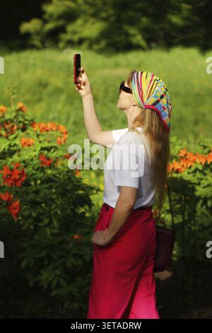Una ragazza scatta foto di fiori in giardino sul suo cellulare in estate. Il blogger di viaggi fotografa la natura, la messa a fuoco selettiva Foto Stock