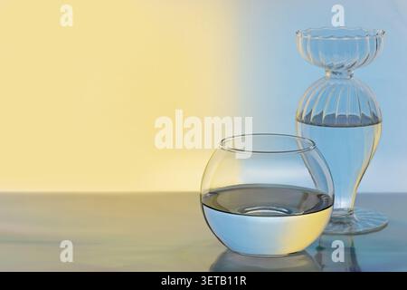 Recipiente in vetro trasparente con acqua e vaso a coste su sfondo giallo pastello blu per un benessere minimo Foto Stock