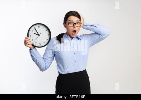 Donna in abbigliamento Business sembra sorpresa con un orologio che indica l'ora tarda Foto Stock