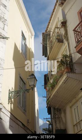 Stretto vicolo di Lisbona con lampada da parete in ghisa ornata, balcone in ferro con piante in vaso e cielo blu sopra Foto Stock
