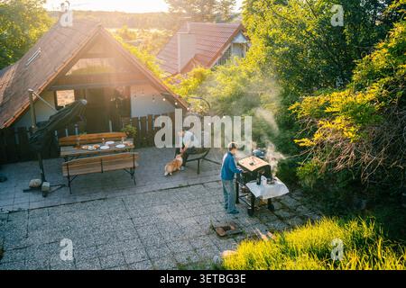 Famiglia che cucina al barbecue fuori da una casa al tramonto con un cane nelle vicinanze e un tavolo apparecchiato per un pasto Foto Stock