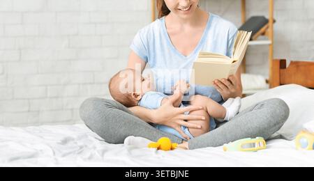 Madre che legge un libro al suo bambino seduto sul letto in una camera accogliente Foto Stock