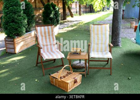 Accogliente area picnic all'aperto con due sedie pieghevoli in legno, cuscini a righe, un tavolino con radio d'epoca e un cestino di vimini su un prato verde. Foto Stock