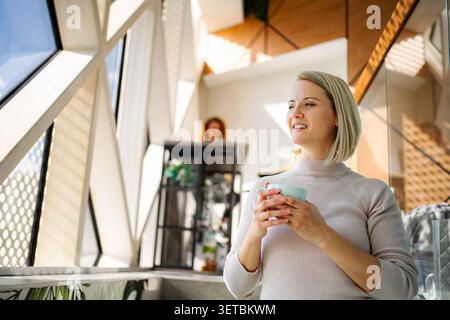 Donna felice che beve caffè guardando lontano godendo di interni moderni Foto Stock