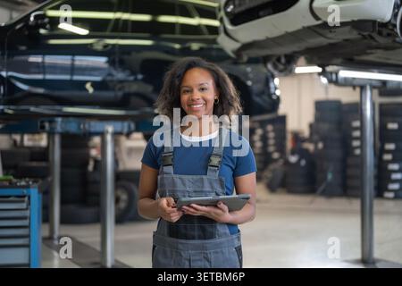 Donna afro-americana sorridente, un meccanico di auto che usa un tablet nel servizio di manutenzione, in piedi accanto ai veicoli su un sollevatore idraulico, ritratto. Foto Stock