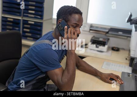 Un meccanico maschio è al telefono con un cliente mentre lavora presso un computer in un'officina di riparazione automobilistica. Foto Stock