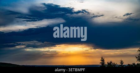 Un tramonto drammatico con raggi dorati di sole che si fanno strada attraverso pesanti nuvole di tempesta. Paesaggio pittoresco con silhouette di alberi all'orizzonte e un contr Foto Stock