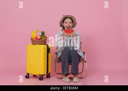 Una donna asiatica felice in un cappello si siede su una sedia da campeggio, tenendo una fetta di anguria davanti al viso, con un cestino di frutta sul suo bagaglio giallo, gustando un sano spuntino estivo. Foto Stock