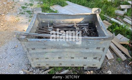 Una cassa di impilamento in acciaio resistente agli agenti atmosferici contenente barre d'armatura filettate e dadi di bloccaggio si trova su un cantiere in ghiaia Foto Stock