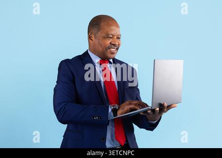 Uomo d'affari afroamericano maturo in costume da marina sorridente mentre digita sul laptop Foto Stock
